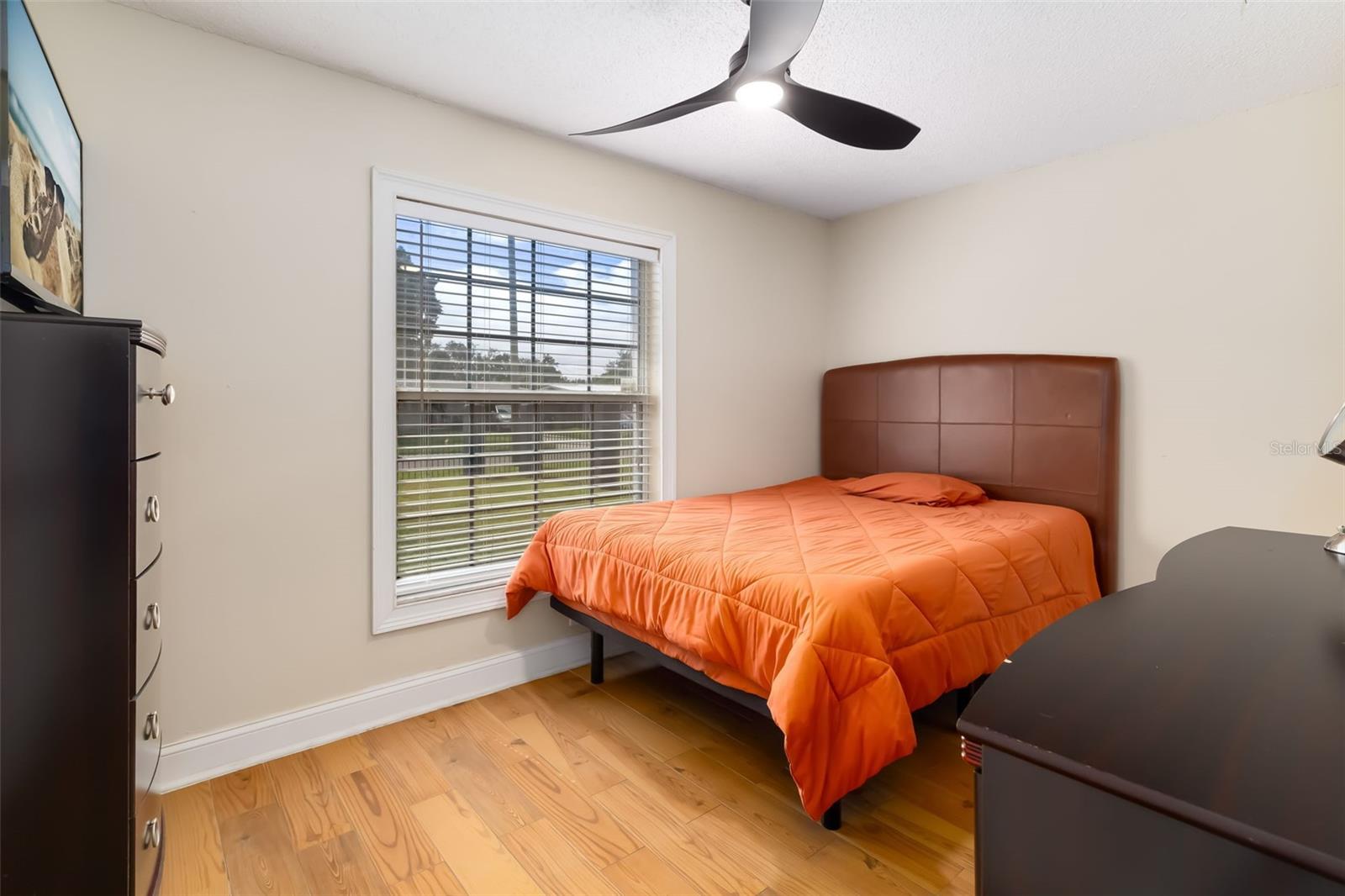 Bedroom with an orange-quilted bed beneath a window with blinds and a ceiling fan.