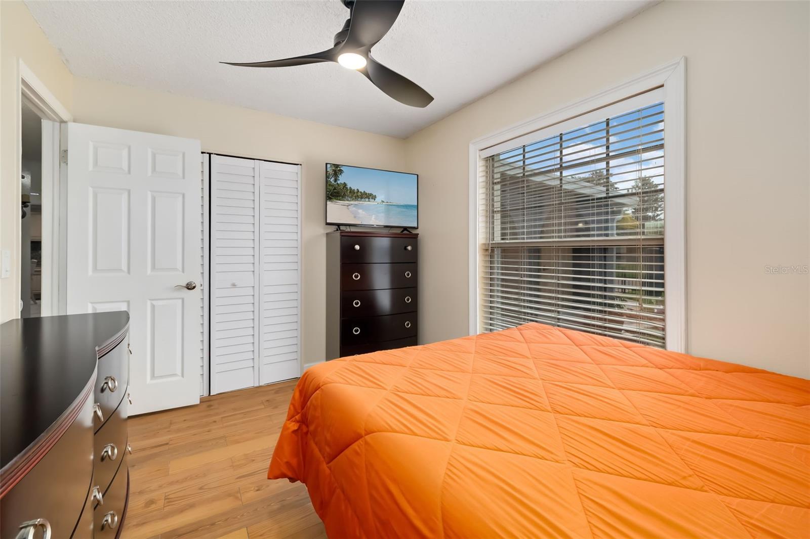 Bedroom with a bright orange quilted bedspread, wood floors, and a large window with blinds.