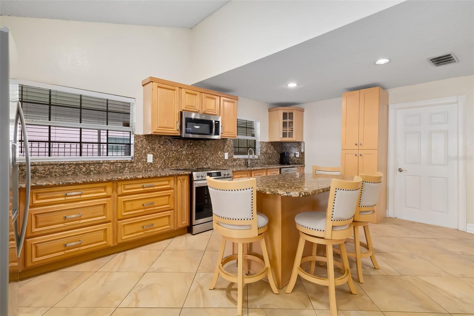 Kitchen with honey-colored cabinets, stone-pattern counters, stainless appliances, and an island with four bar stools.