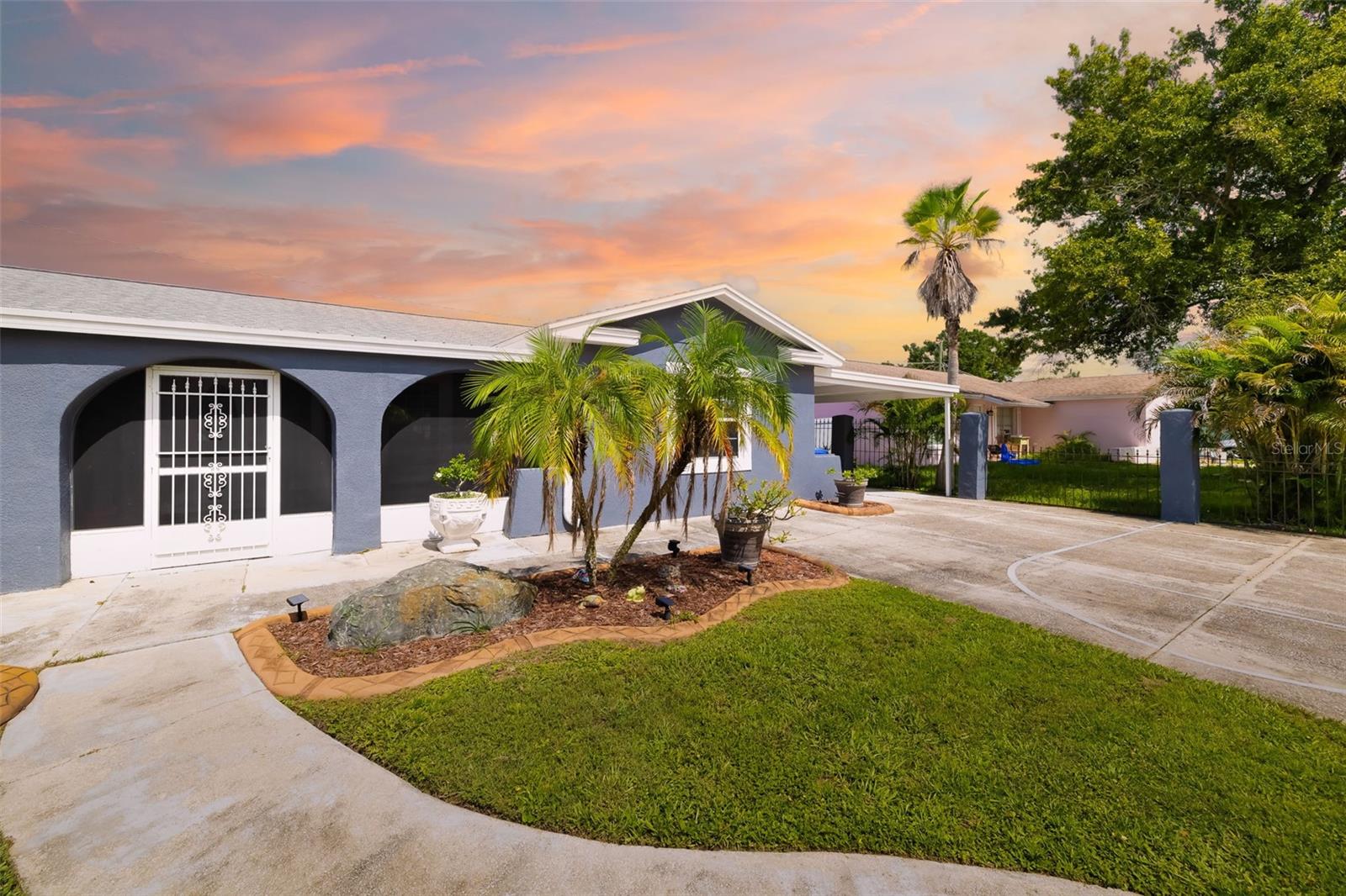 Single-story gray stucco home with arched entryways, tropical palms, and a curved driveway at sunset.