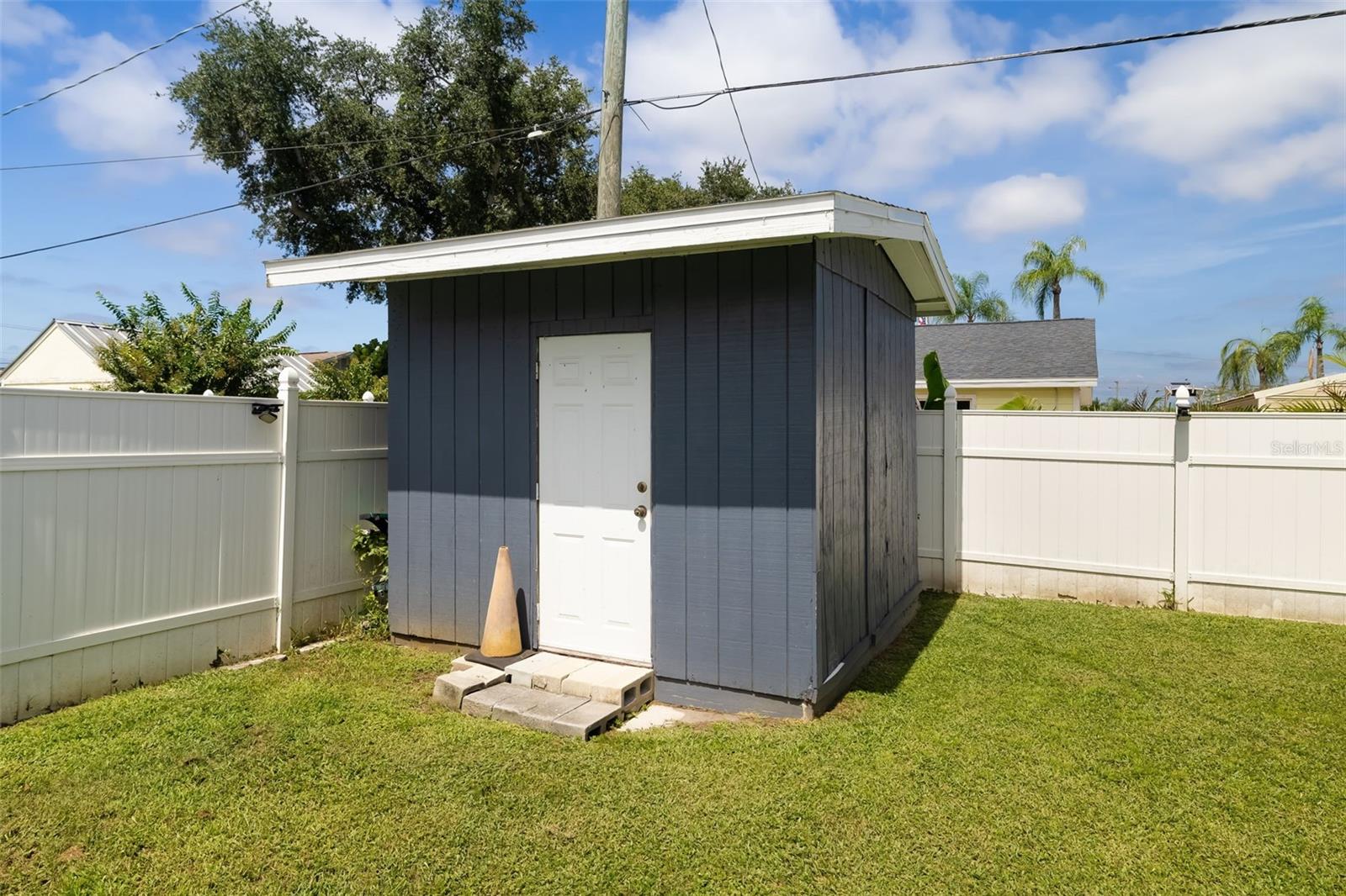 Dark blue backyard shed with a white door and sloped roof inside a fenced lawn.