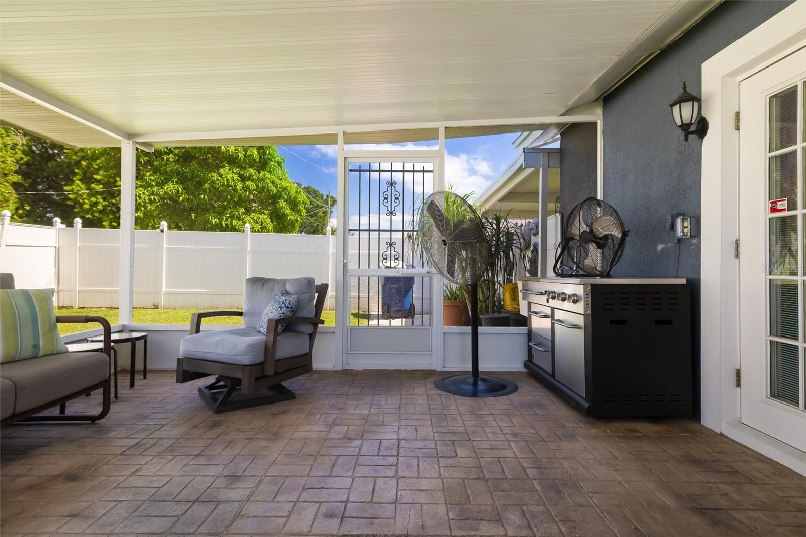 Covered patio with cushioned seating and a stainless-steel grill beside a fenced yard.