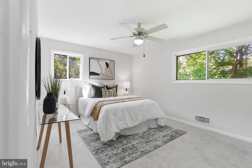Staged bedroom with a white bed, ceiling fan, and windows overlooking leafy trees.