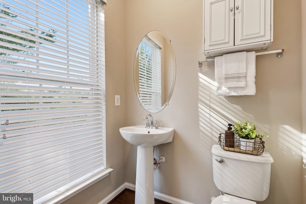 Bathroom with a pedestal sink and oval mirror beside a sunlit window with white blinds.
