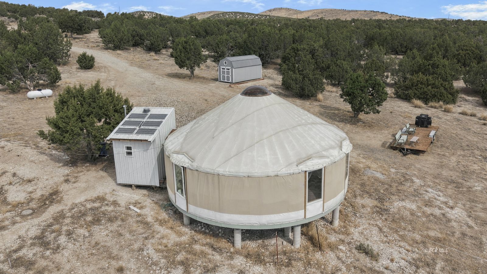 Round yurt-style home on a wooded rural lot with solar panels and outbuildings.