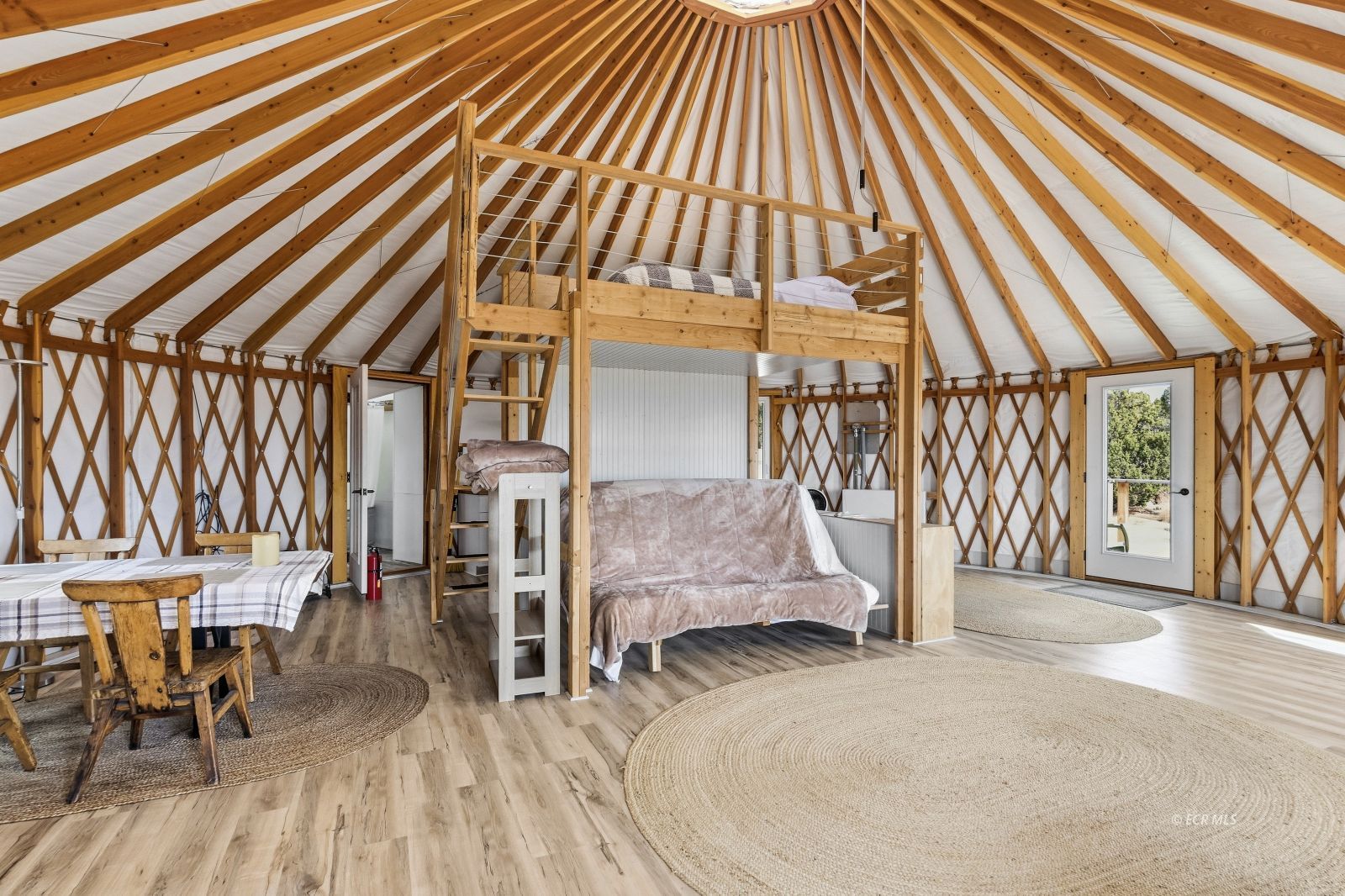 Yurt interior with exposed timber rafters, a loft bed, sofa, and dining table.