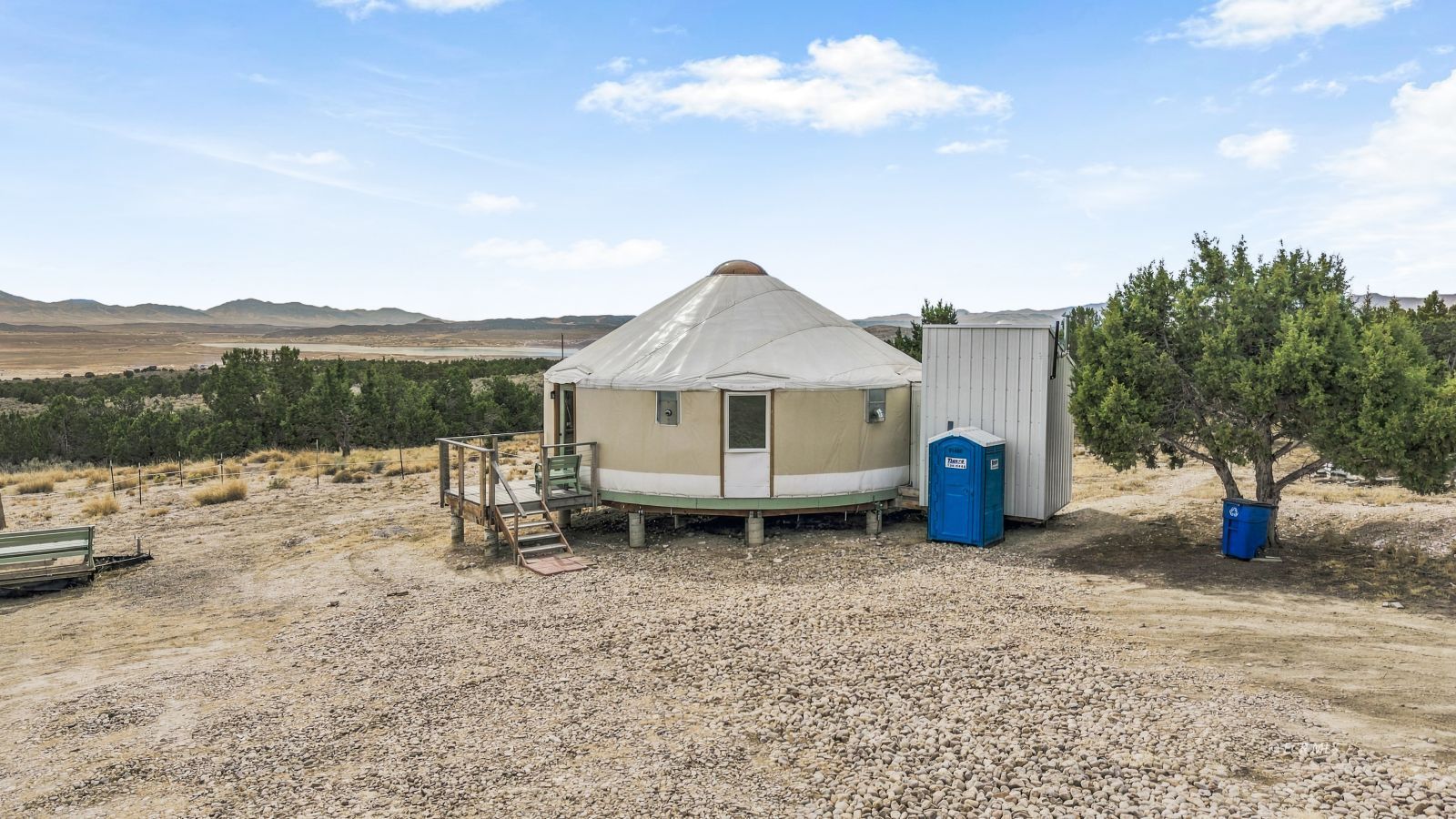 Round beige yurt with a white conical roof on a rocky rural lot beneath distant mountains.