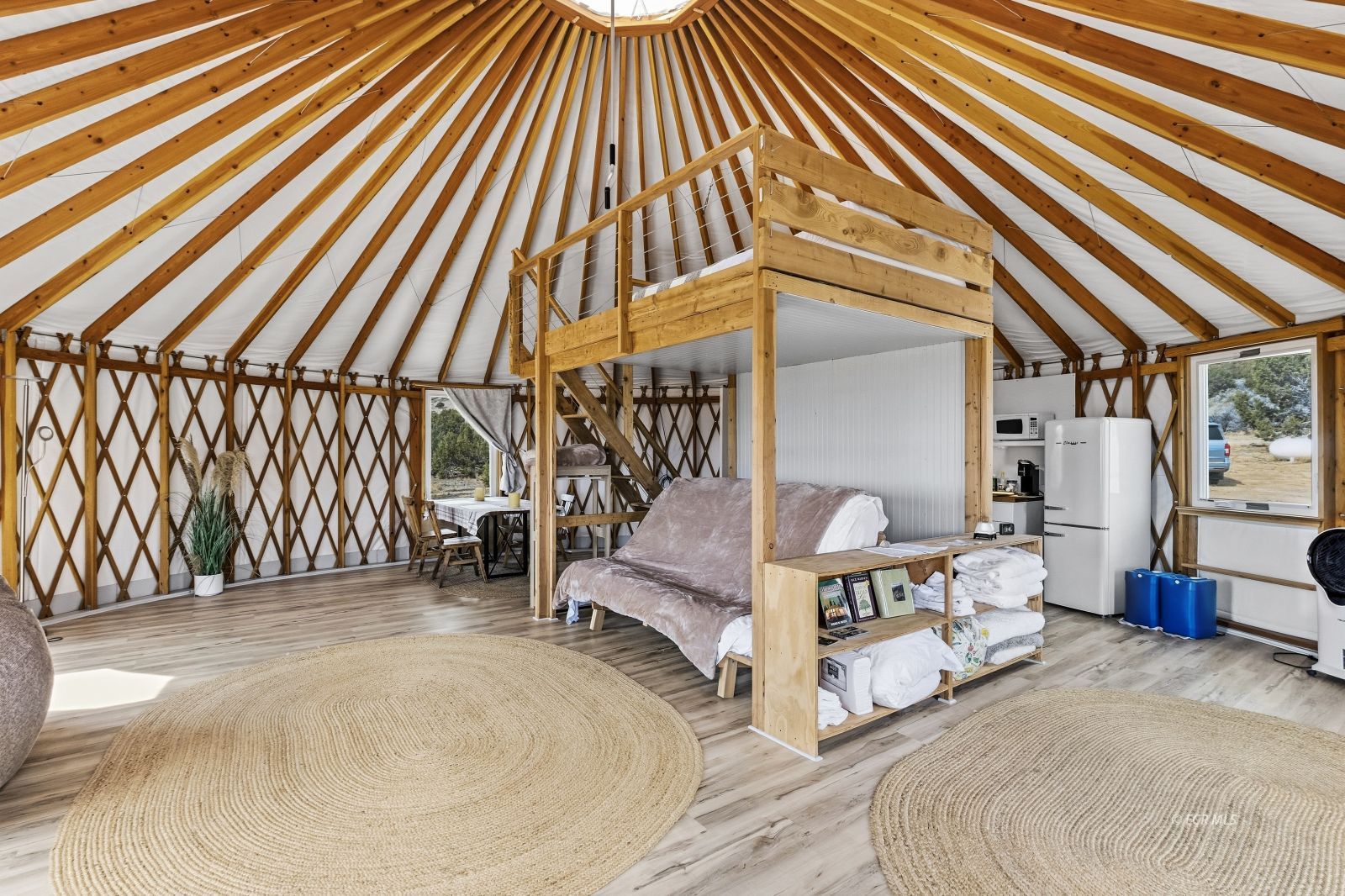 Furnished yurt interior with a loft bed, kitchenette, and exposed radial timber rafters.