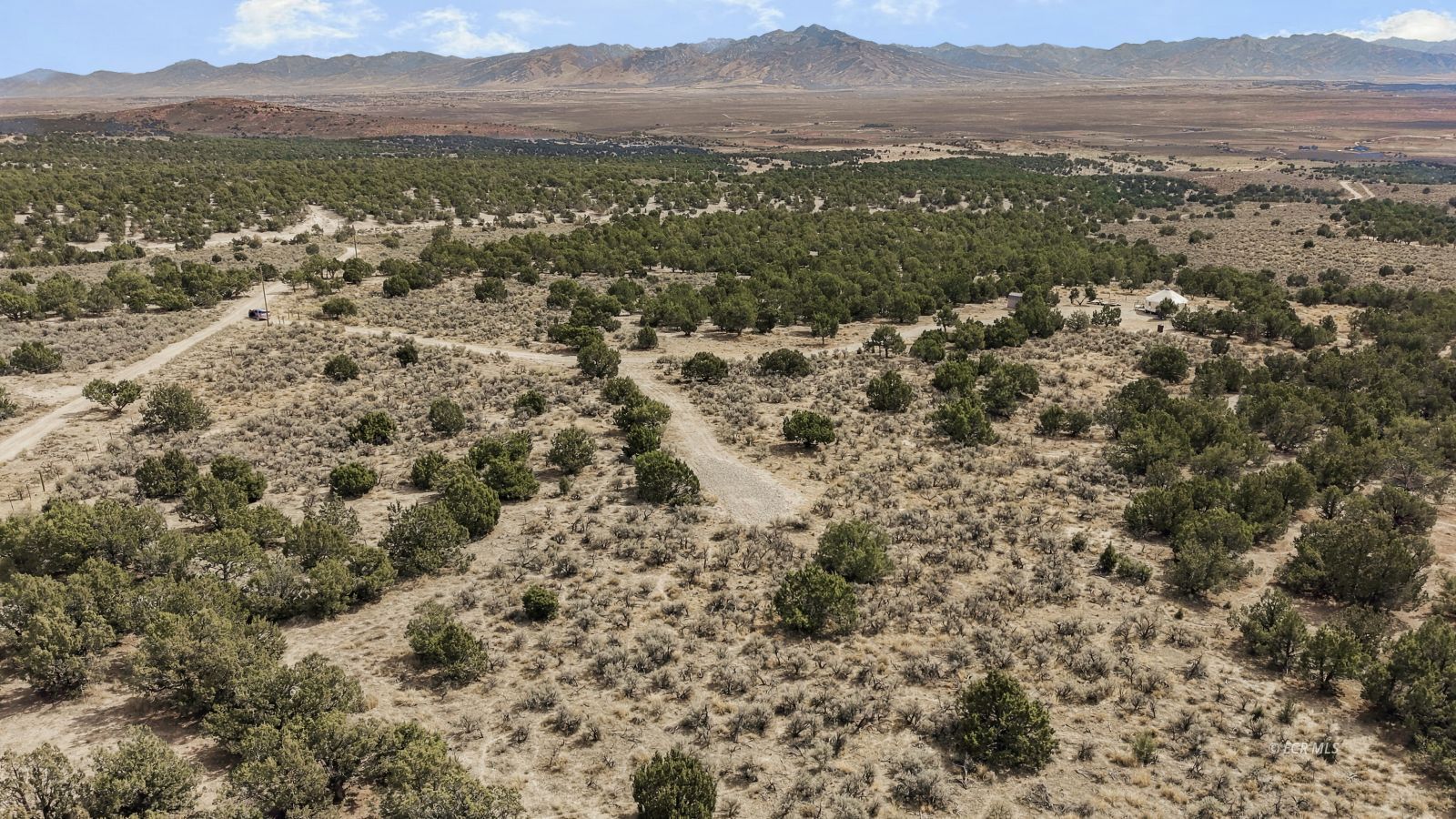 Aerial view of open land with scattered juniper, dirt tracks, and distant mountains.