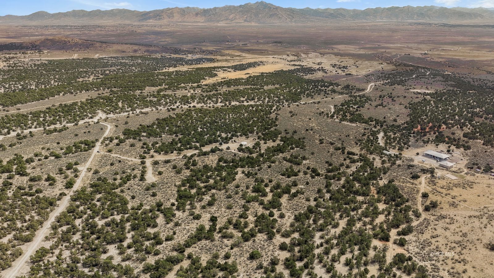 Aerial view of scrub-covered acreage with dirt tracks, a small building, and distant mountains.