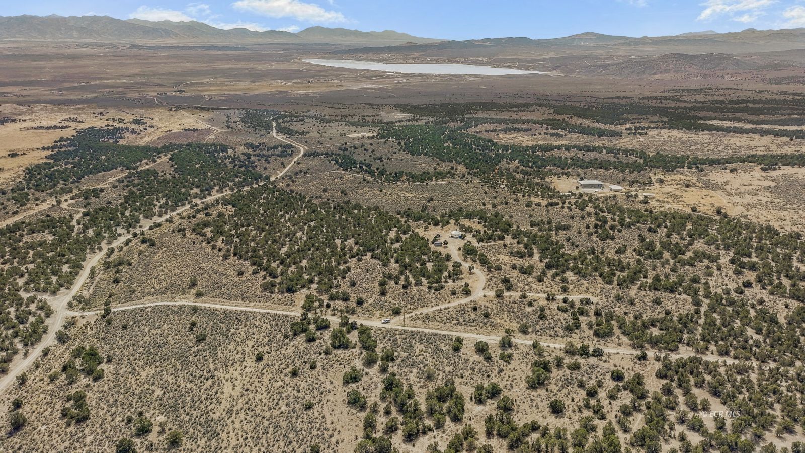 Aerial view of rural desert land with winding dirt roads, scattered buildings, a reservoir, and distant mountains.