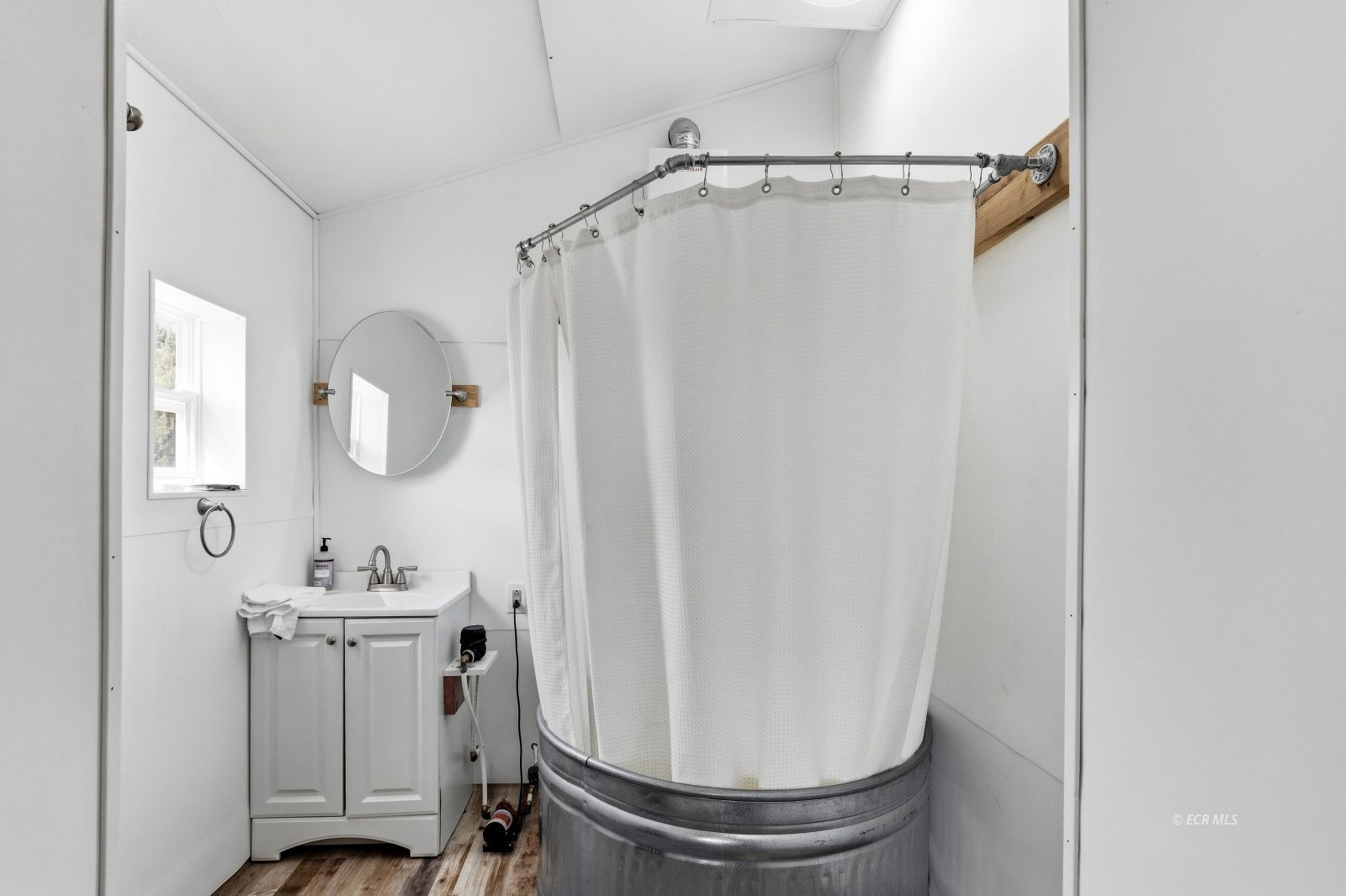 Bathroom with a round galvanized-metal shower enclosure, white curtain, and small vanity beneath a window.