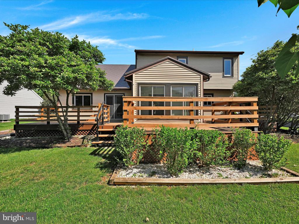 House rear with a broad wooden deck, lawn, shrubs, and mature trees.