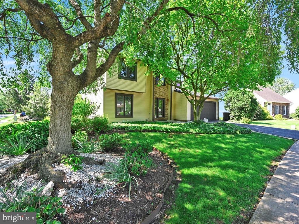 Two-story yellow-sided home framed by mature trees, with landscaped garden beds and a green front lawn.