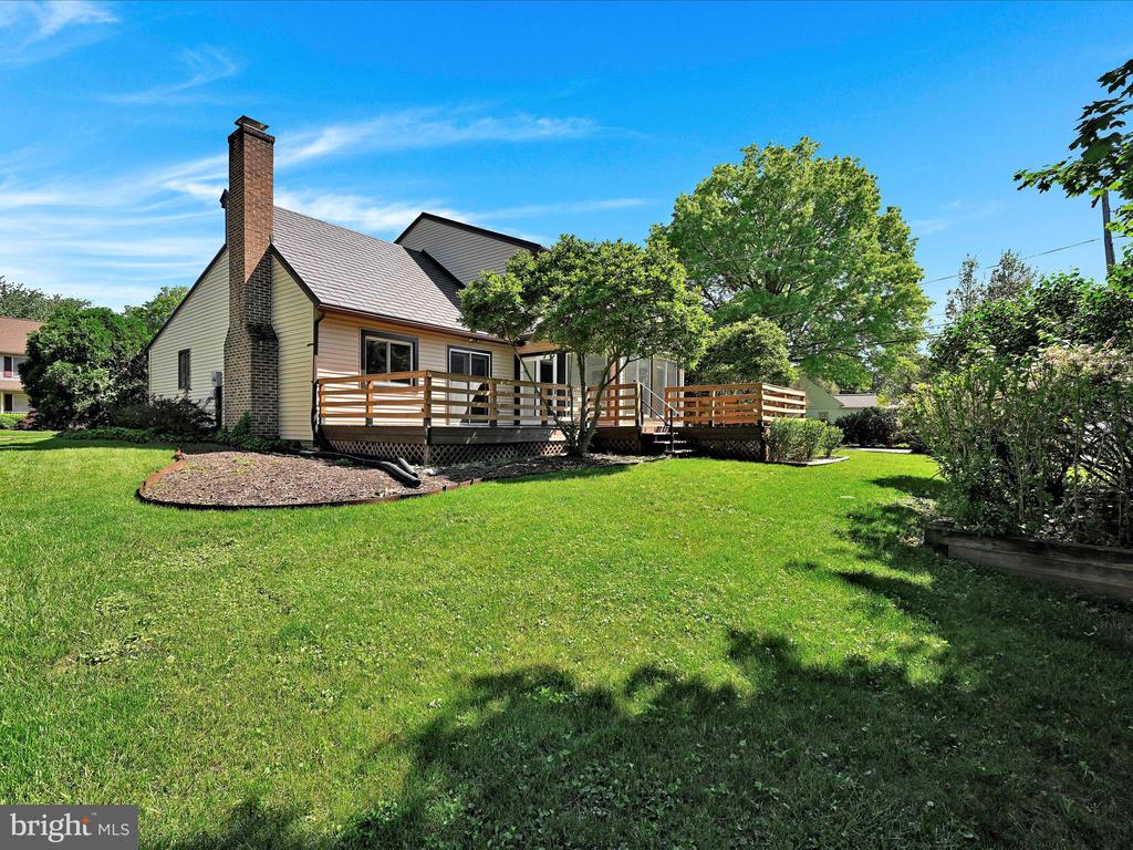 Rear of a light-colored house with a brick chimney and raised wooden deck overlooking a broad lawn.