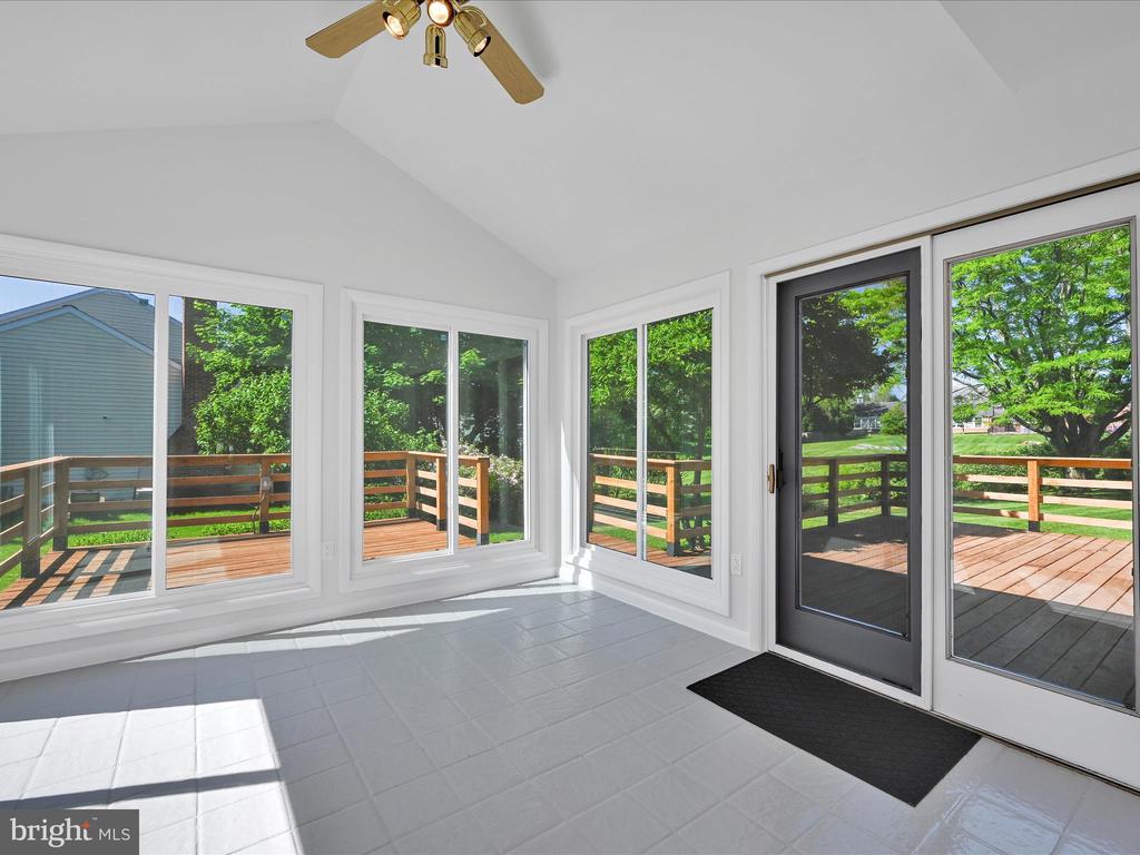 Sunroom with a vaulted ceiling, white tile floor, and large windows overlooking a wooden deck and leafy yard.