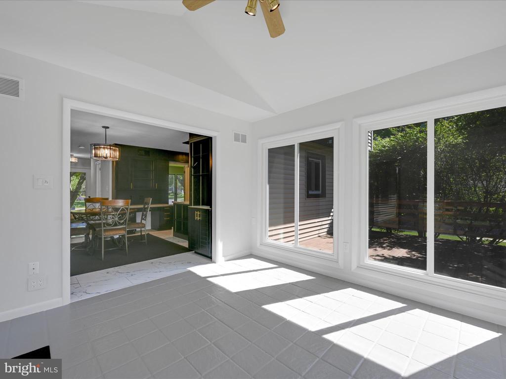Sunroom with large windows overlooking a shaded deck, opening to a dining area and green kitchen.
