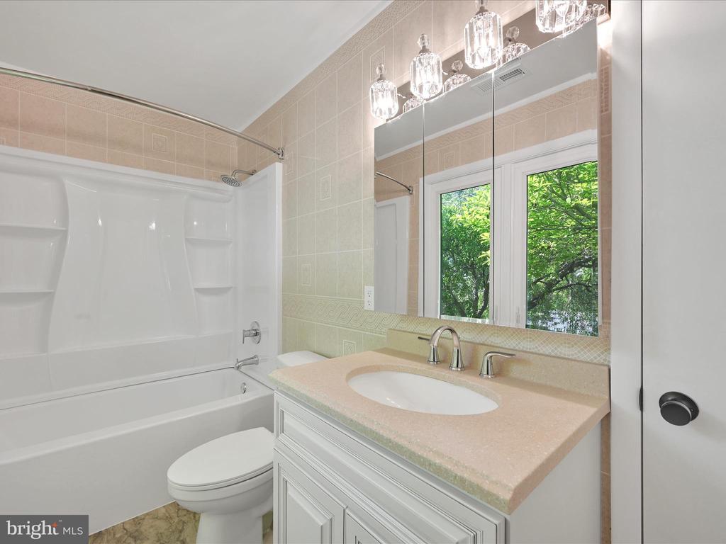 Bathroom with a white tub and shower, pale stone vanity, and mirrored cabinet reflecting leafy windows.