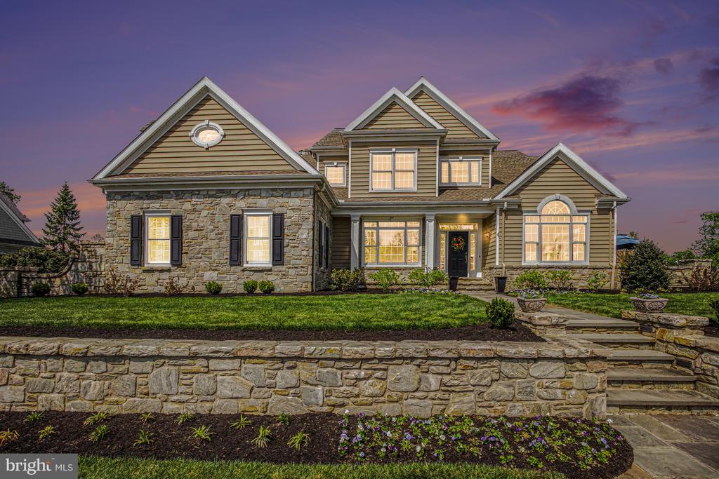 Large stone-and-siding home with illuminated windows, landscaped lawn, and a stone retaining wall at dusk.
