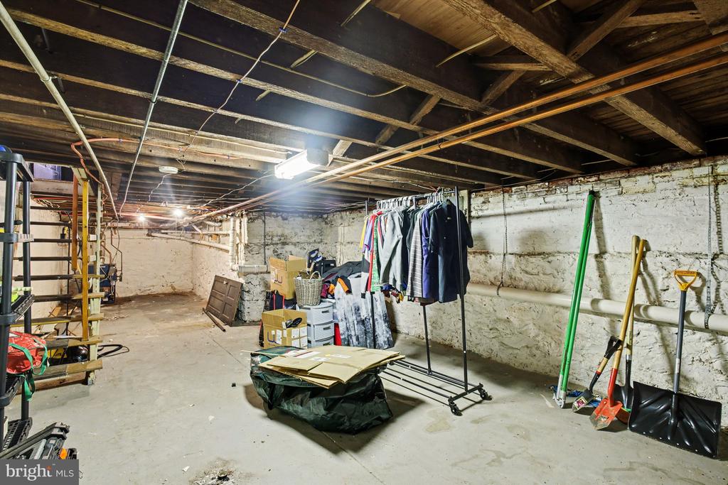 Basement with exposed joists, stone walls, utility pipes, and a clothes rack.