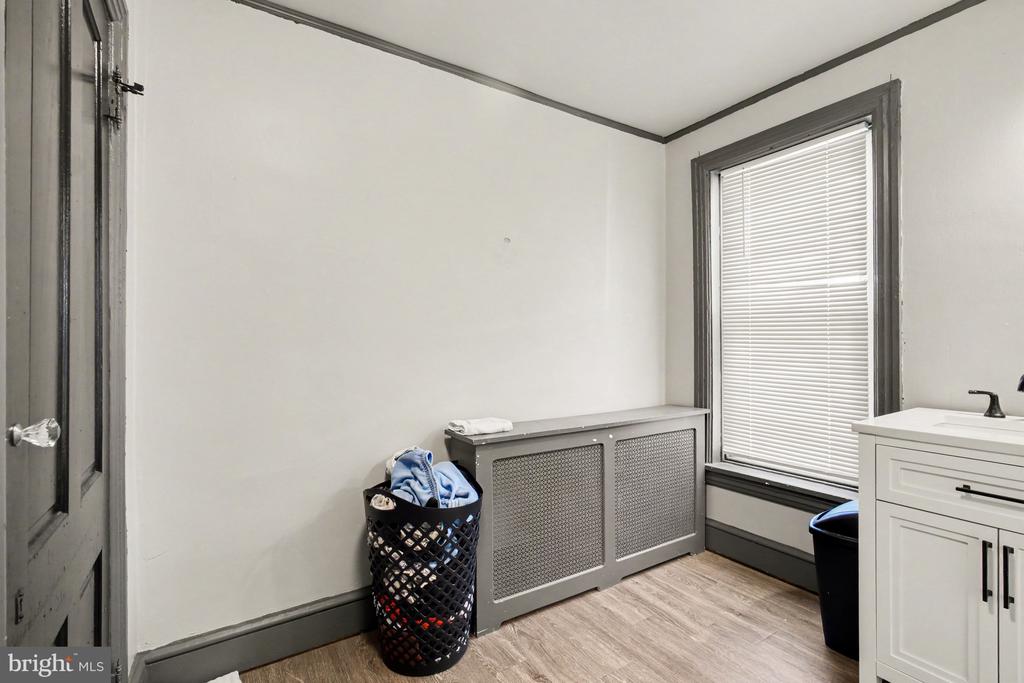 Bathroom with a white vanity, window blinds, and covered radiator beside a laundry basket.