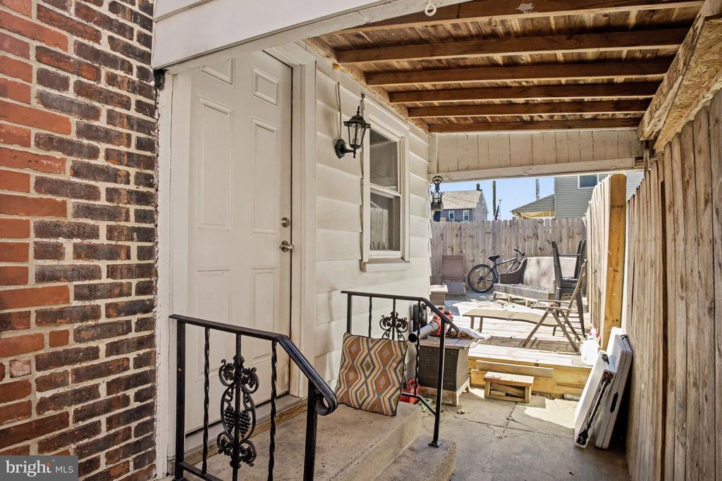 Covered patio with a white side entrance, brick wall, and fenced outdoor seating area.