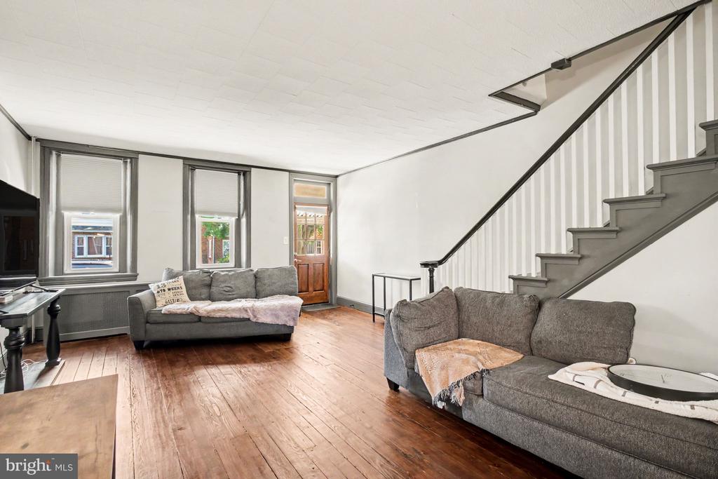 Living room with hardwood floors, gray sofas, tall windows, and a staircase.