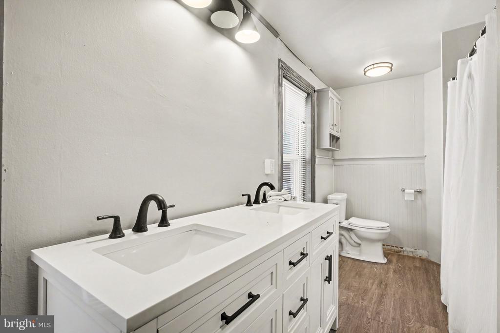 Bathroom with a double-sink vanity, dark fixtures, toilet, and wood-look flooring.