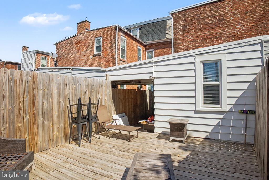 Fenced wooden deck patio beside a white-sided home, with outdoor furniture and brick row houses behind it.