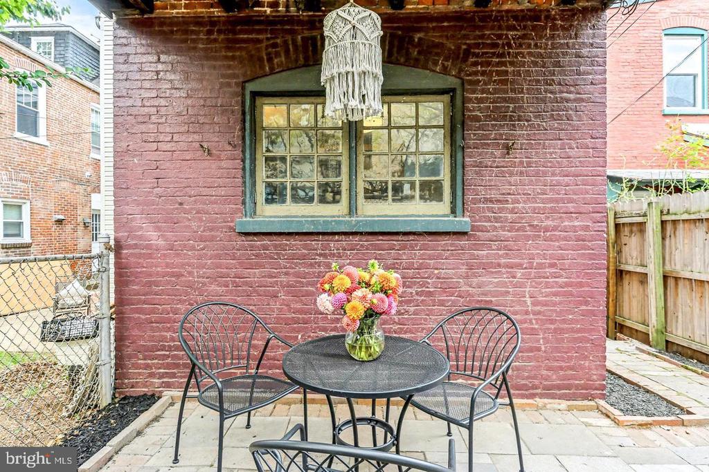 Brick-walled patio with a black metal table, chairs, and colorful flowers.