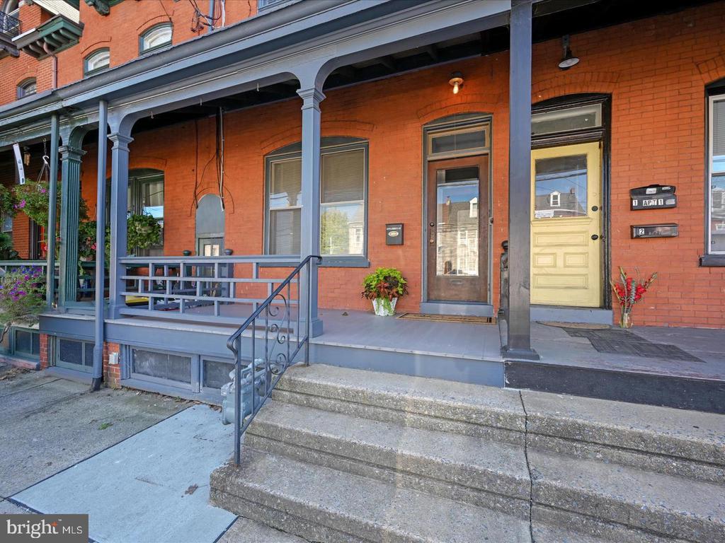 Red-brick building with a covered porch, blue-gray railings, concrete steps, and brown and yellow entrance doors.