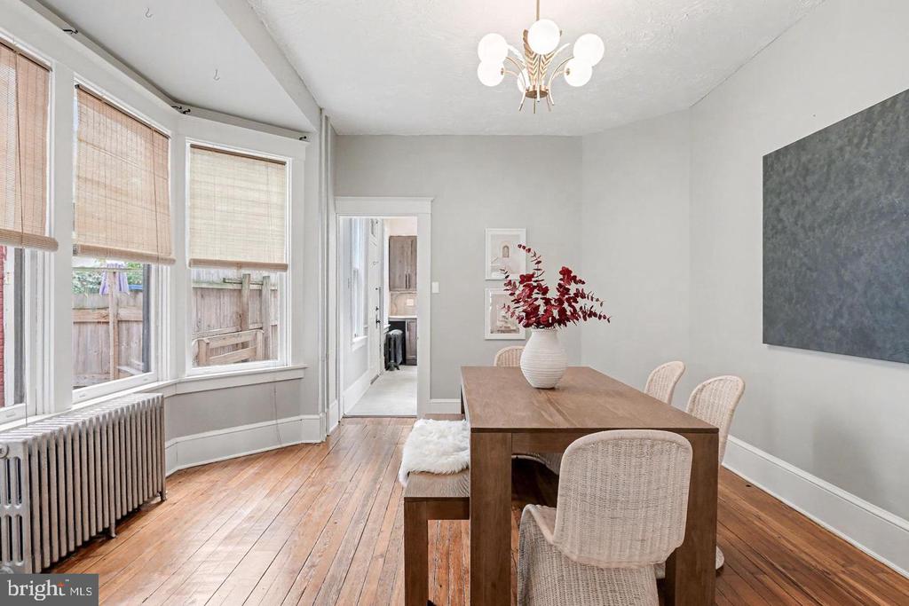 Dining room with a wooden table, woven chairs, tall shaded windows, and hardwood floors.