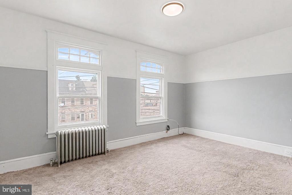 Empty carpeted room with two tall windows and a radiator beneath one.