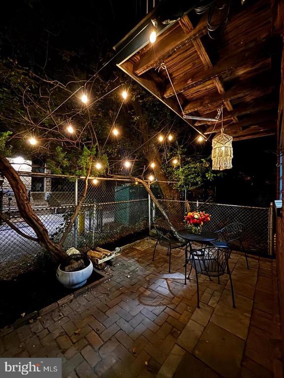 Covered brick patio with string lights, outdoor dining set, and potted tree at night.