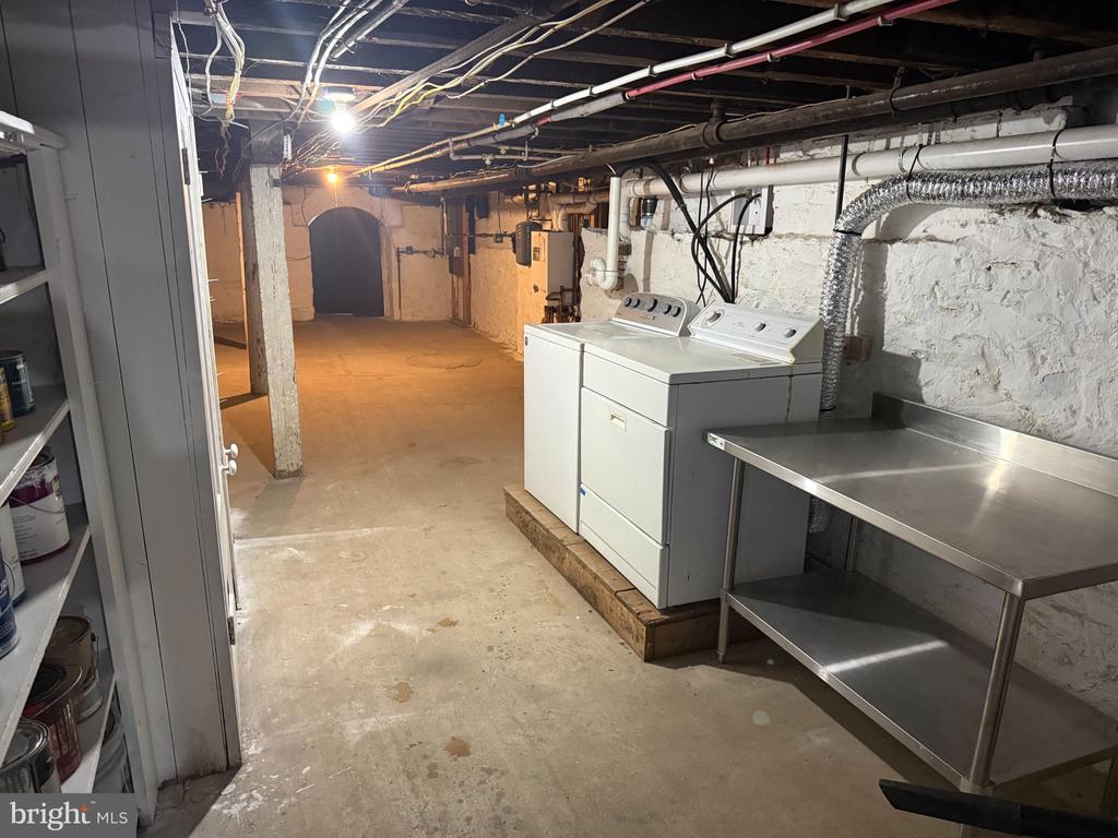 Basement laundry area with washer, dryer, stainless-steel worktable, and exposed ceiling pipes.