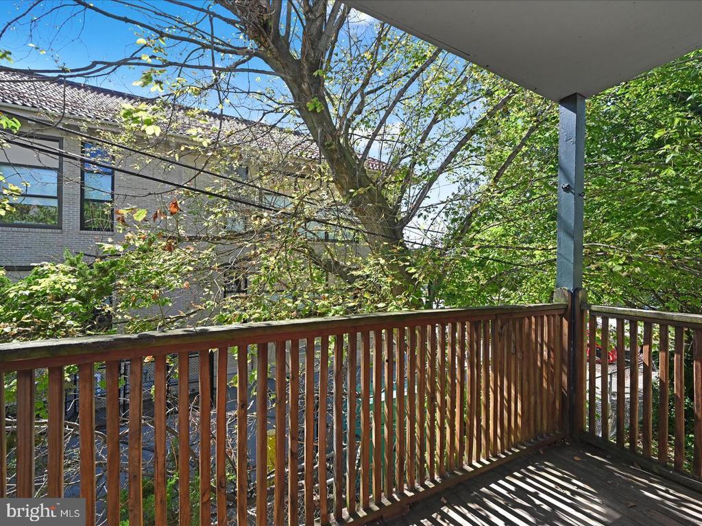 Covered wooden deck with brown railing overlooking leafy trees and a neighboring brick building.