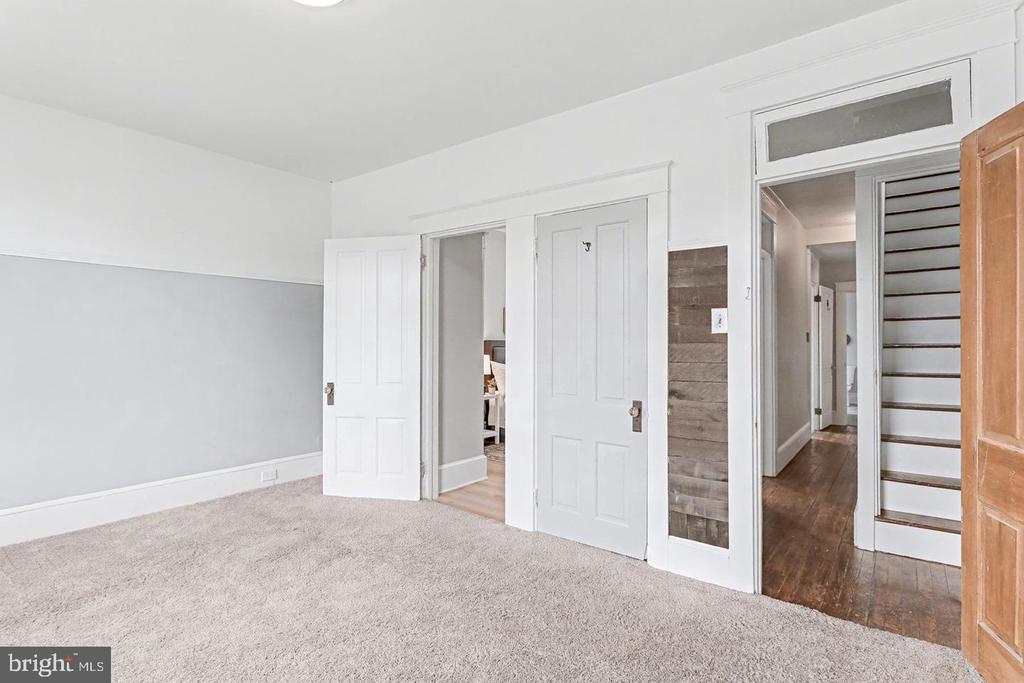 Vacant carpeted room with white paneled doors and a staircase visible through the hallway.