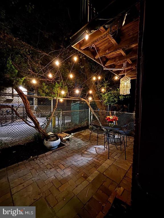 Nighttime patio with brick pavers, outdoor dining set, string lights, and a fenced yard.