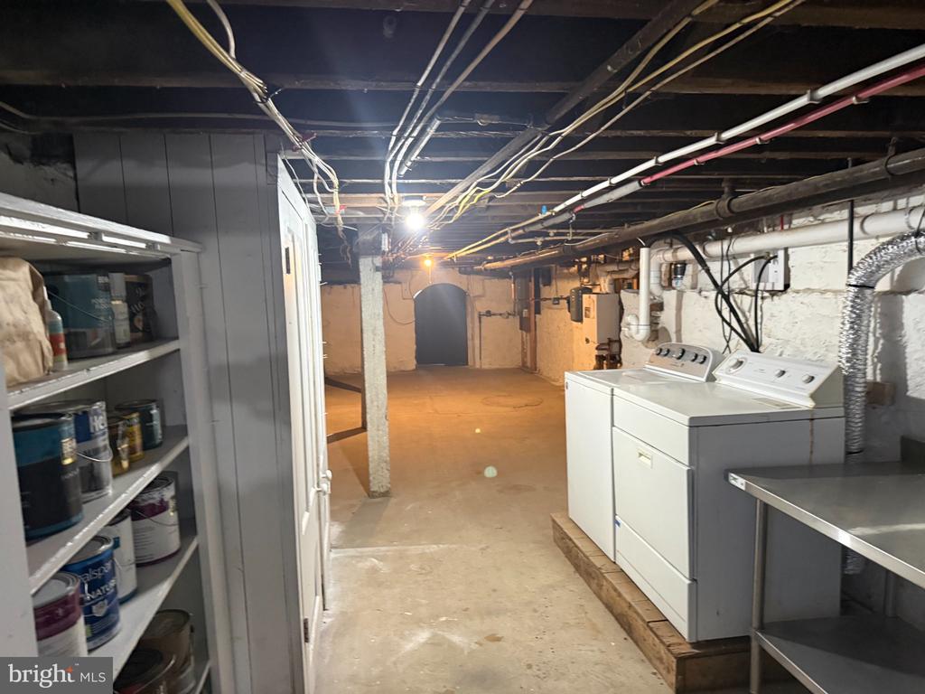 Basement laundry area with a washer, dryer, exposed pipes, and paint cans on shelves.