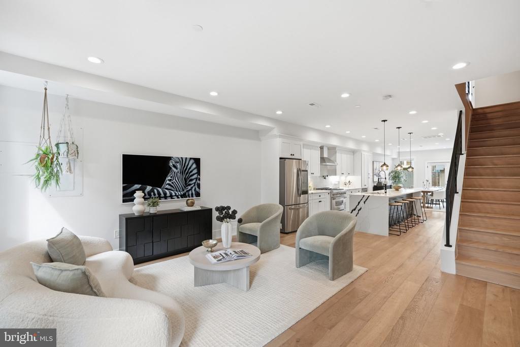 Open-plan living room and kitchen with light seating, white cabinetry, and a wood staircase.