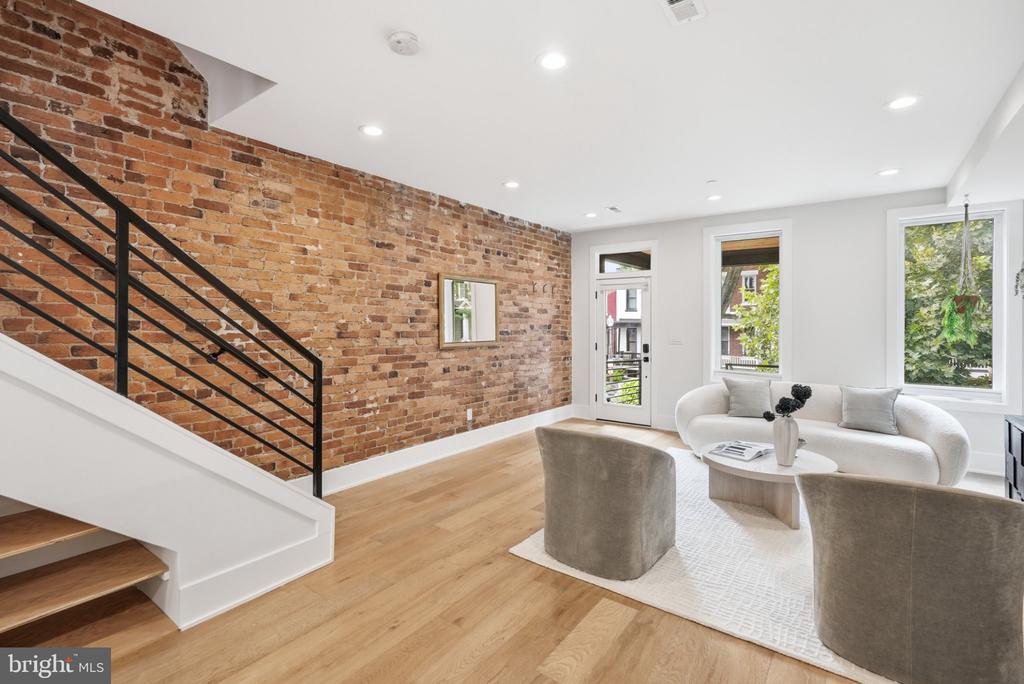 Living room with an exposed brick wall, black-railed staircase, and white sofa by tall windows.