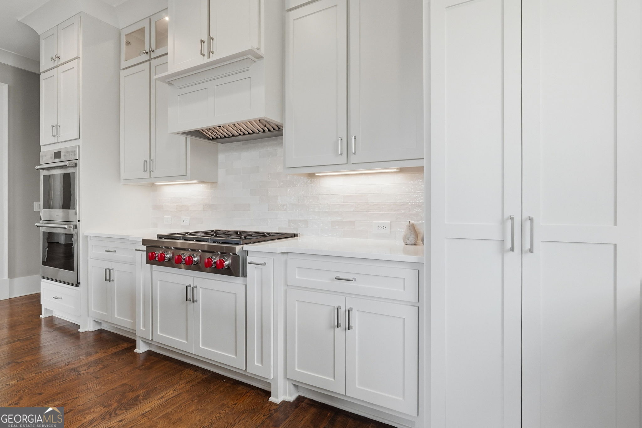 White kitchen with shaker cabinets, stainless double oven, gas range with red knobs, and dark wood flooring.