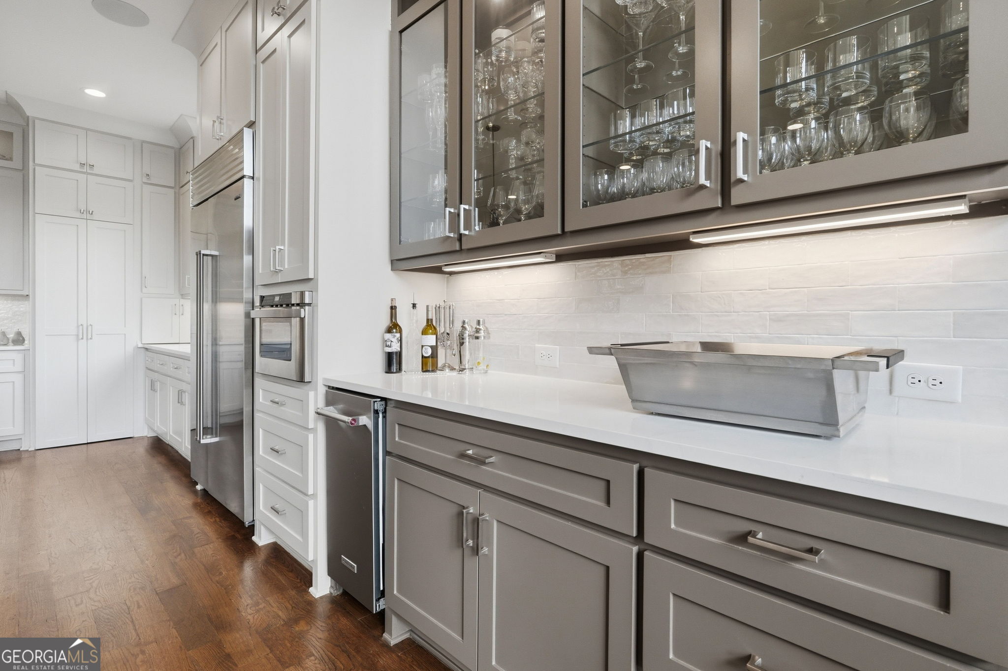 Modern kitchen with glass-front cabinets, stainless steel appliances, and a long white countertop.