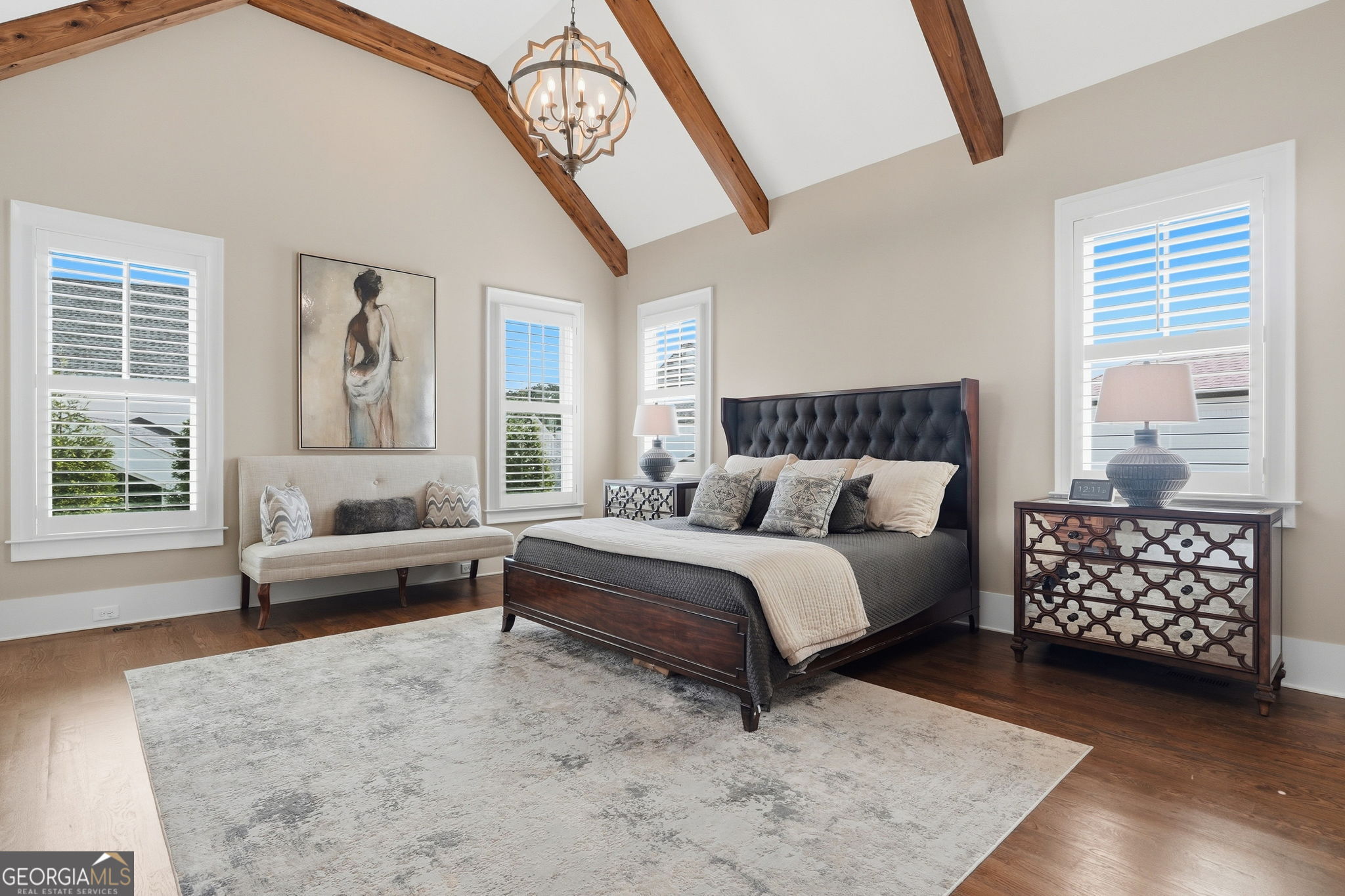 Staged bedroom with exposed wood ceiling beams, a dark tufted bed, and plantation-shuttered windows.