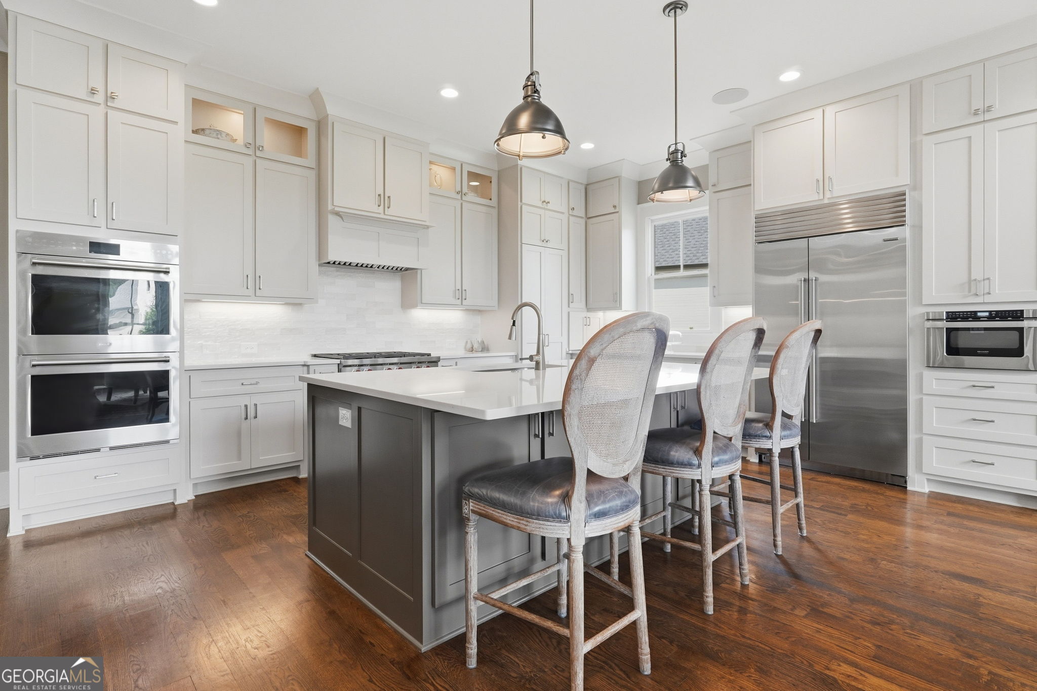 Kitchen with white cabinetry, dark island, three bar stools, stainless-steel appliances, and hardwood floors.