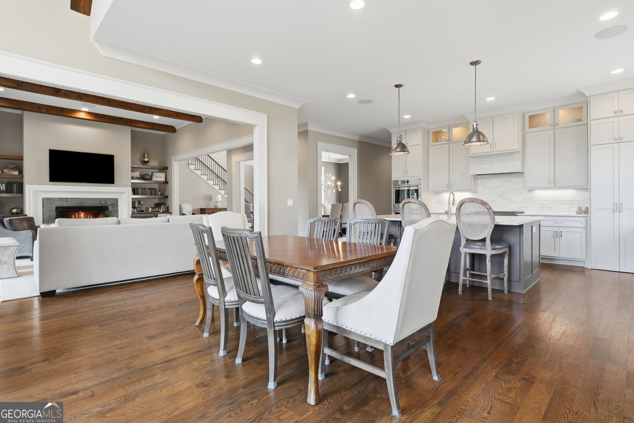 Open-plan dining room with a carved wood table, white kitchen cabinetry, hardwood floors, and an adjoining fireplace.
