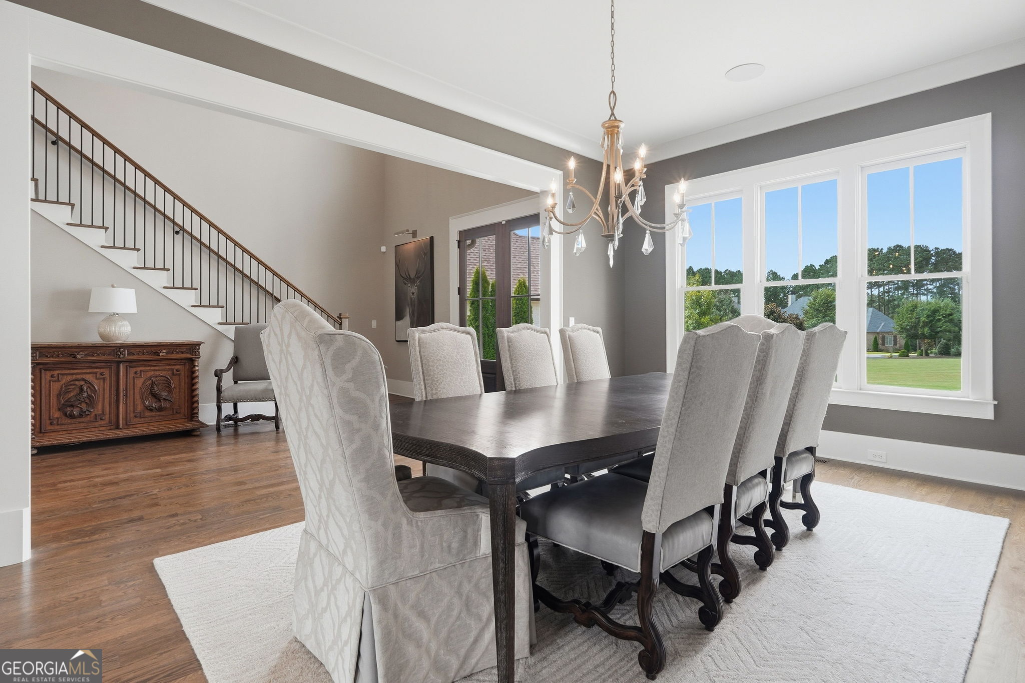 Dining room with a dark wood table, upholstered chairs, chandelier, and staircase beside large windows.