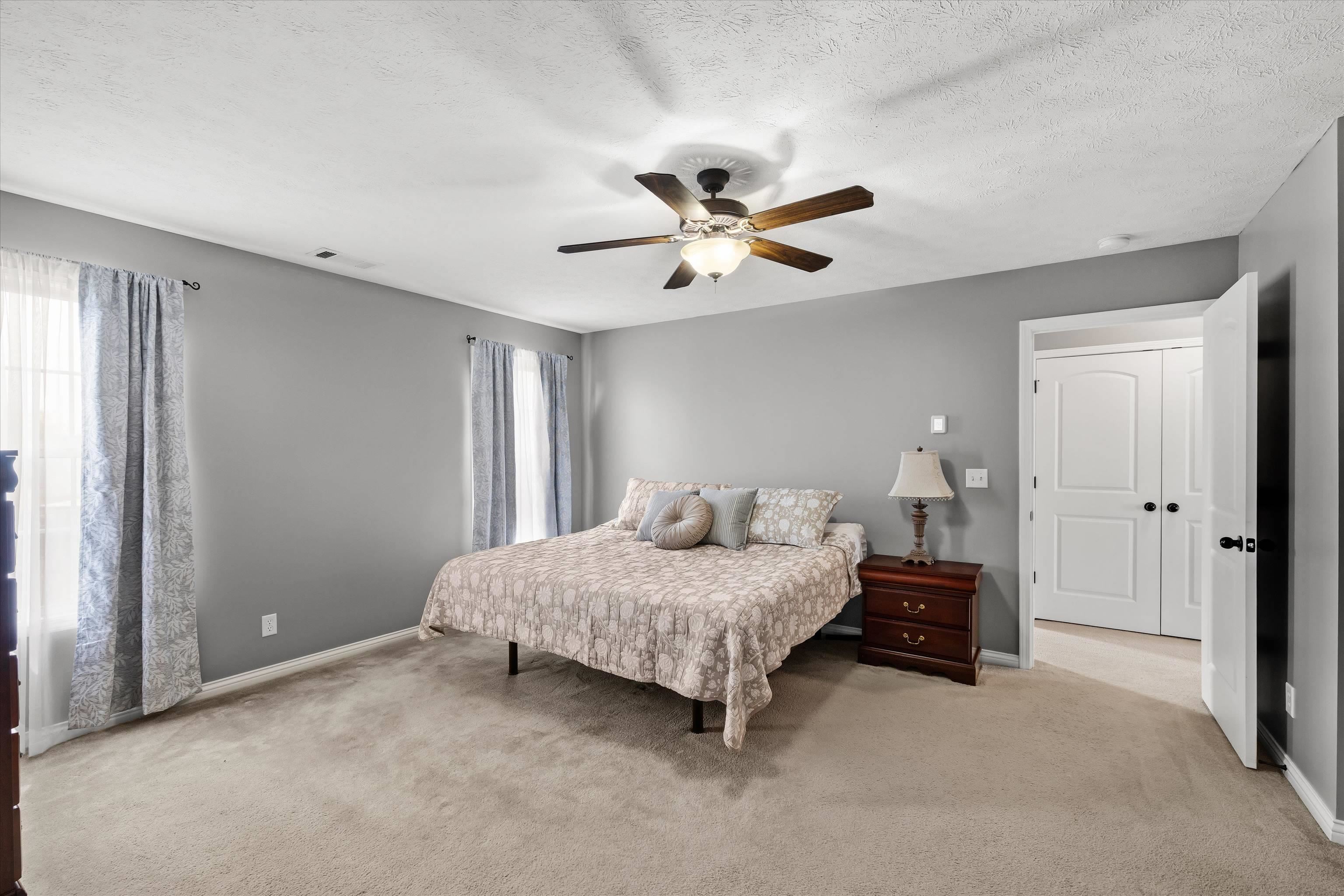 Furnished bedroom with gray walls, beige carpet, patterned bedding, and a ceiling fan with light.