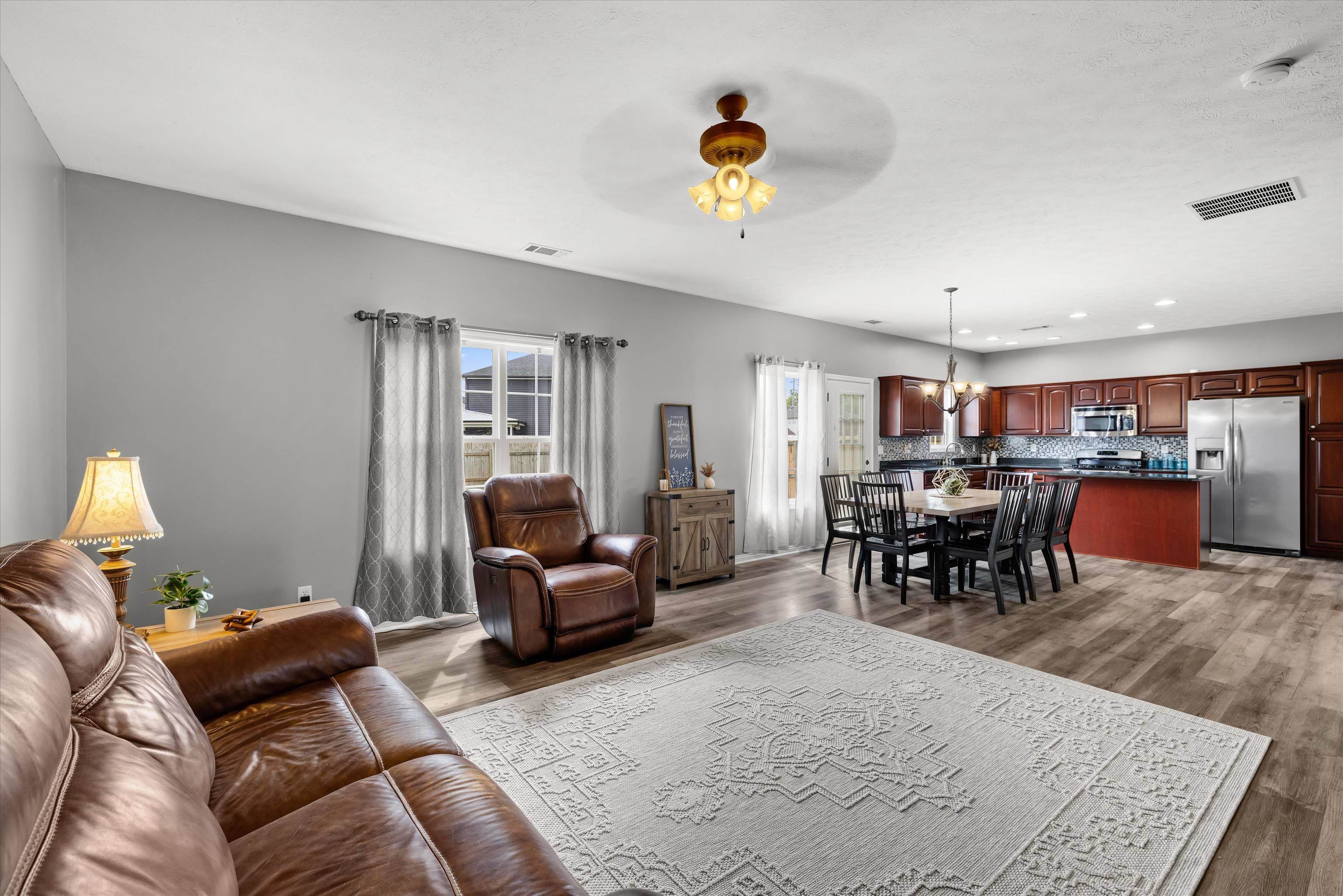 Open-plan living and dining area with brown leather seating, patterned rug, and dark wood kitchen cabinets.