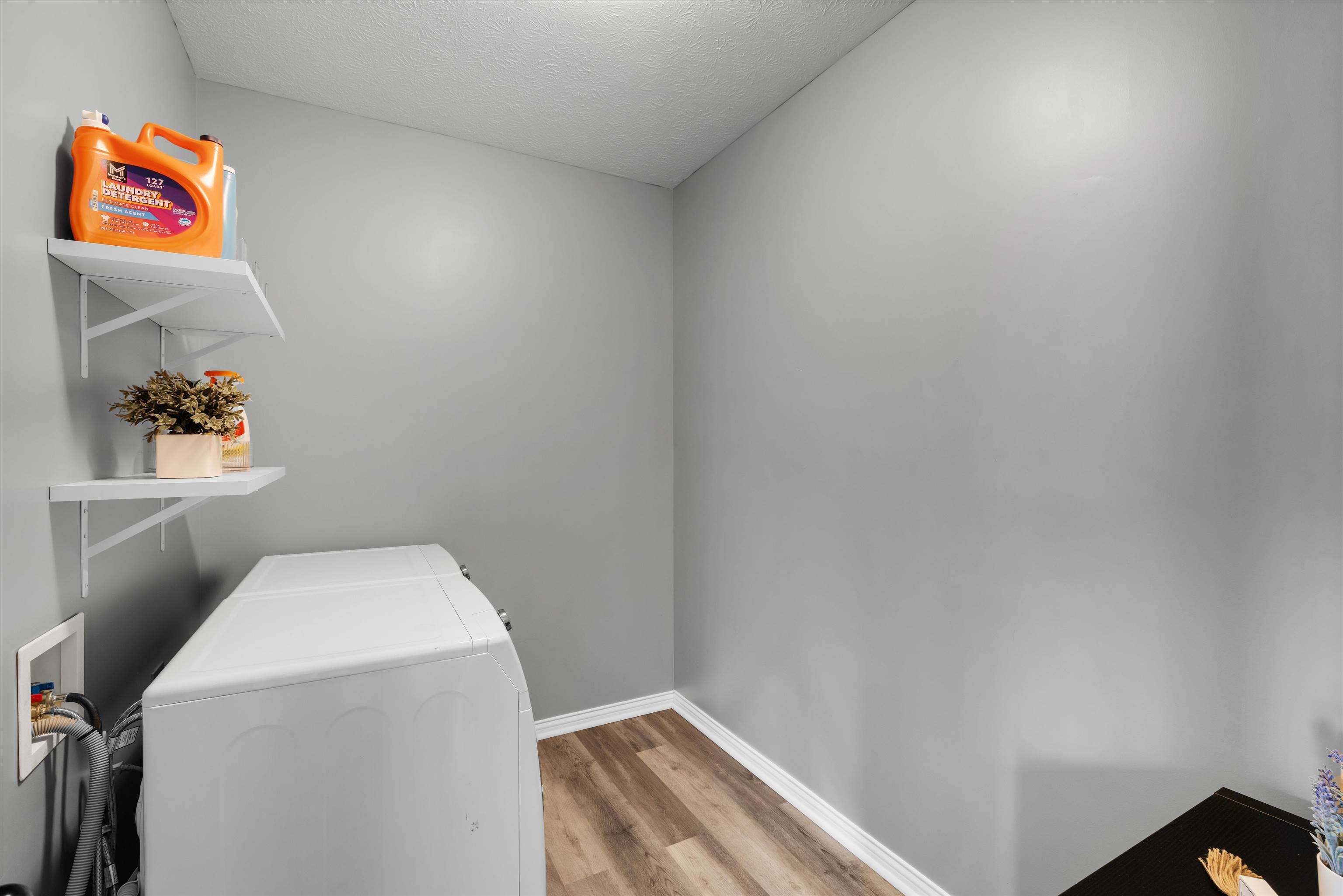 Laundry room with a white top-loading washer, gray walls, and shelves holding detergent and a small plant.